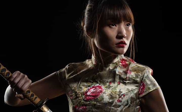 Portrait of a Japanese woman in traditional clothing, holding a katana, captured dramatically against a black background.