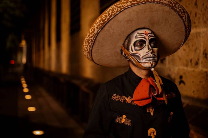 A man in traditional Mexican attire with skull face paint stands in an illuminated street, perhaps during a cultural festival at night.