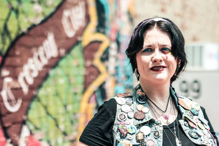 Portrait of a woman in denim vest with colorful pins against a vibrant mural background.