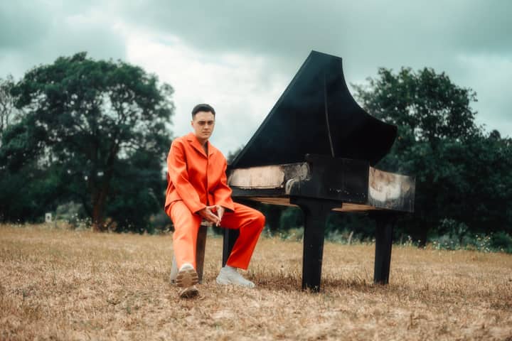 A man in a vibrant orange suit sits by a grand piano in an open meadow.