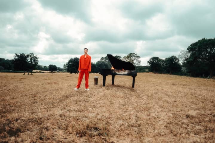 Man in vibrant outfit with flaming piano in a summer meadow.