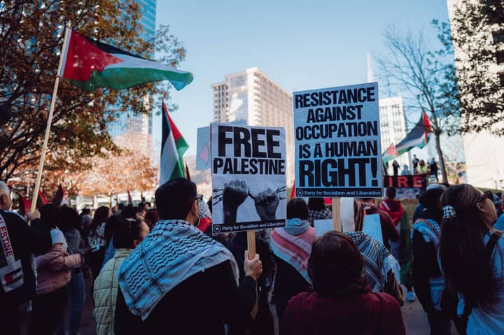 A city rally with protesters holding signs promoting Palestinian rights and human rights advocacy.