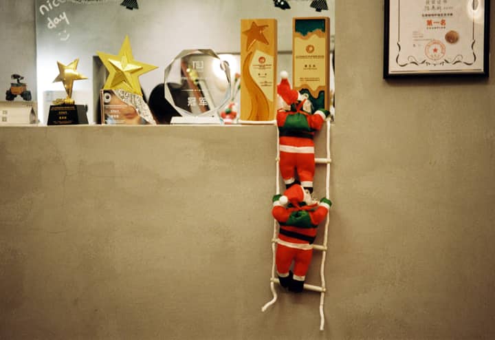 Santa Claus decorations climbing a ladder in a festive office setting, surrounded by awards and certificates.