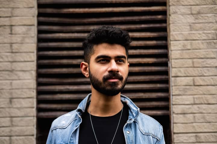 A young bearded man with dark hair wearing a denim jacket, standing outdoors against a textured brick wall.