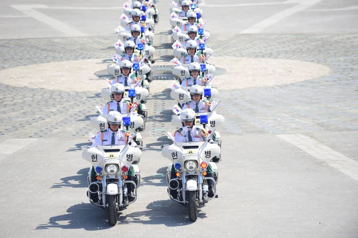A line of South Korean police officers on motorcycles holding flags in an urban parade.