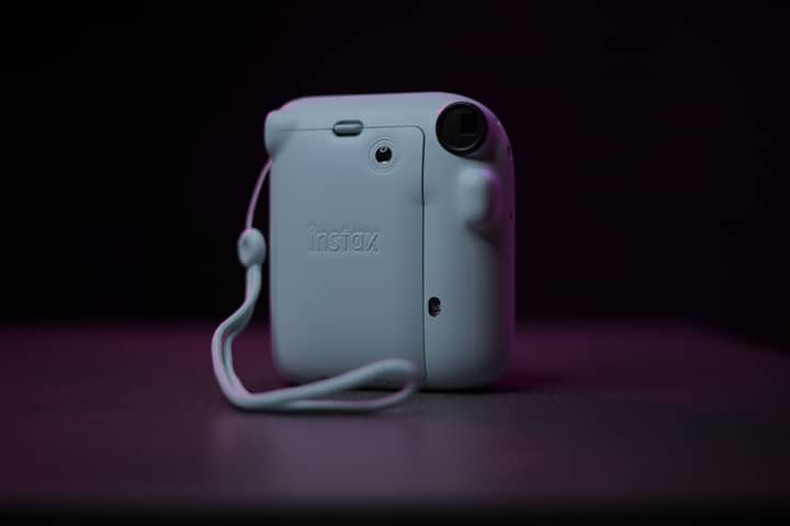 A close-up shot of a Fujifilm Instax Mini camera with a wrist strap, set against a dark background.