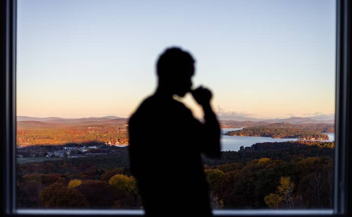 A silhouette of a person drinking by a window with a scenic sunset view over mountains and a lake.