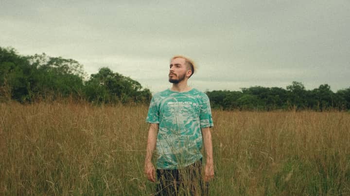 A man with bleached hair stands in a lush rural field, conveying serenity and freedom.