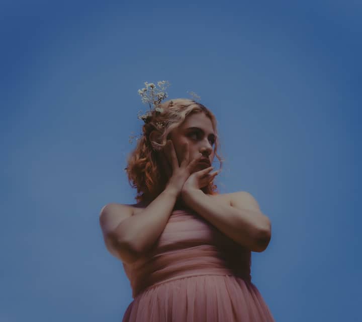 Fashion portrait of a woman in a pink dress against a vibrant clear sky, featuring a floral headpiece.
