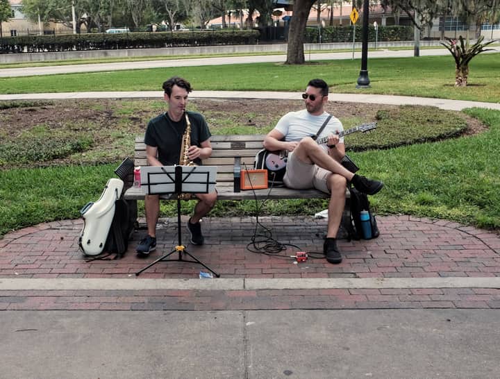 Two musicians playing guitar and saxophone on a park bench in Orlando, FL.