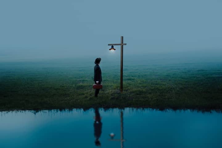 A solitary figure stands under a light post by water, reflecting solitude and hope.
