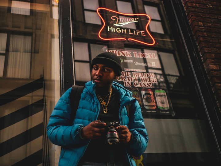 Urban portrait of a man holding a camera, standing by neon signs and window reflections.