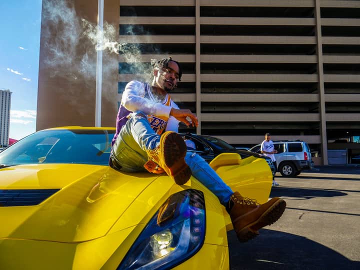 A fashionable man sits on a yellow sportscar in an urban setting, exuding style and confidence.