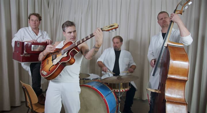 Group of musicians playing guitar, drums, and double bass indoors.