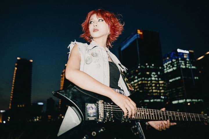 A woman with dyed hair plays an electric guitar against a city night skyline, showing urban rock vibes.