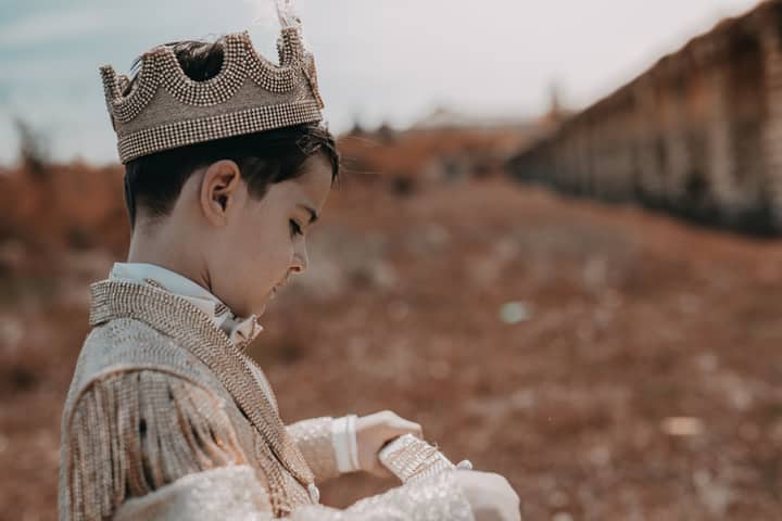 A child dressed as a prince wearing a crown in an outdoor setting with fall scenery.