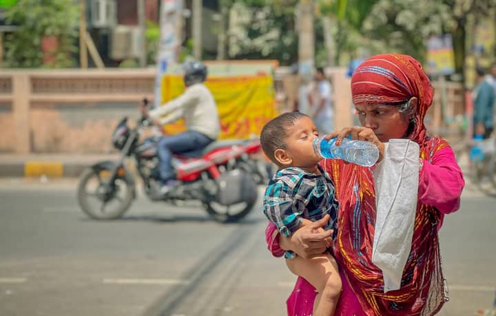 A mother gives water to her child on a hot day in Dhaka, Bangladesh, showing care amidst city life.