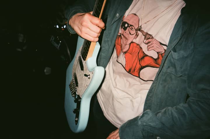 A musician strums an electric guitar in a dimly lit room, wearing a graphic t-shirt.