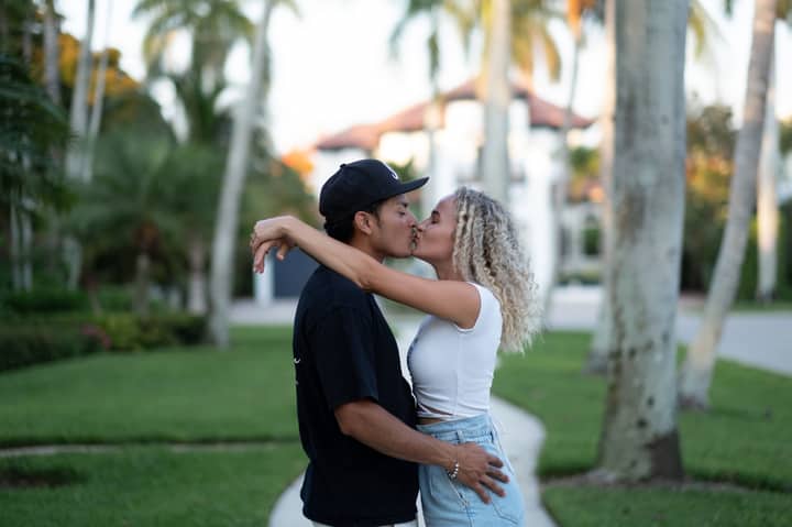 A young couple embraces and shares a romantic kiss on a lush green suburban walkway.