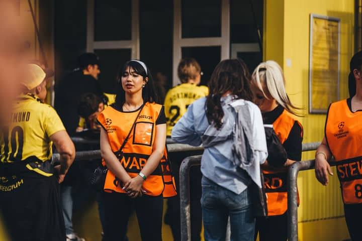 A lively scene with soccer fans and stewards interacting outside a stadium on a sunny day.