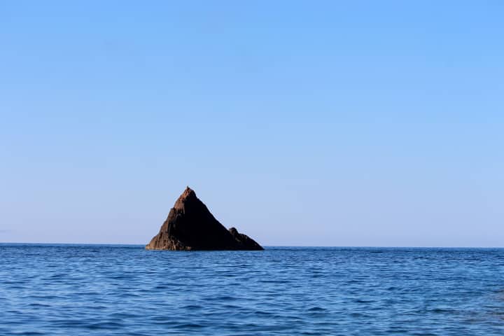 A lone rocky island surrounded by a clear blue ocean under a bright sky.