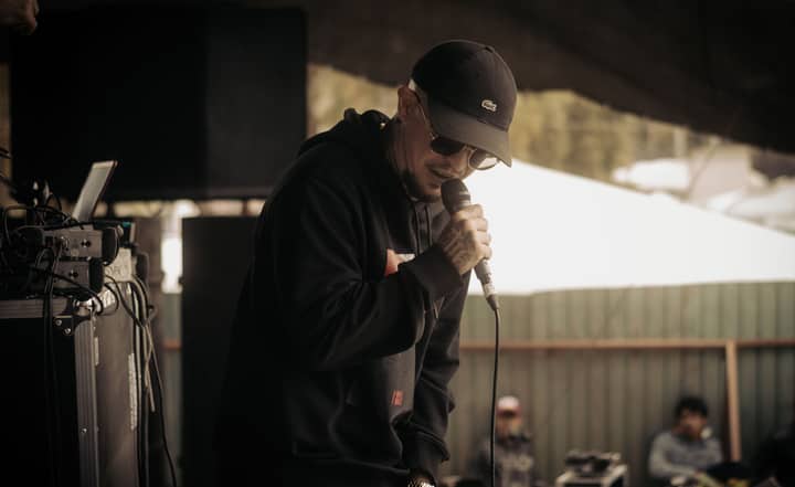 A male singer in a black hoodie performs outdoors with a microphone.