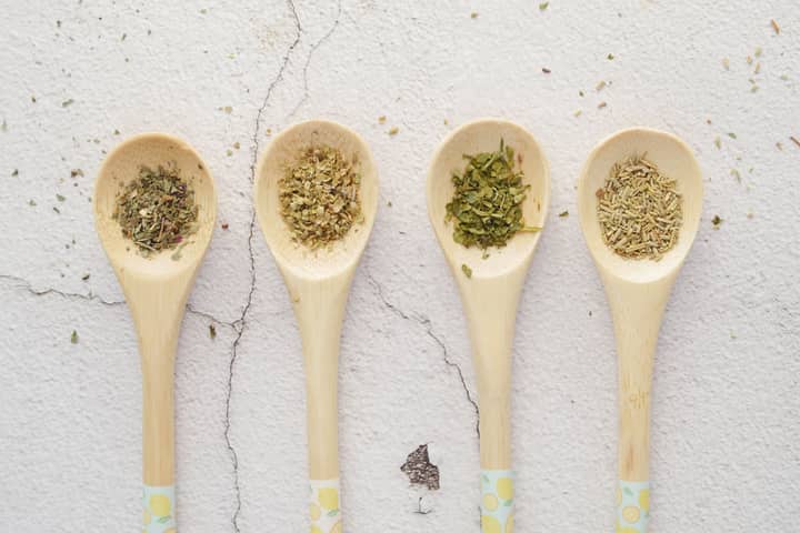 Four wooden spoons with assorted dried herbs on a light background.