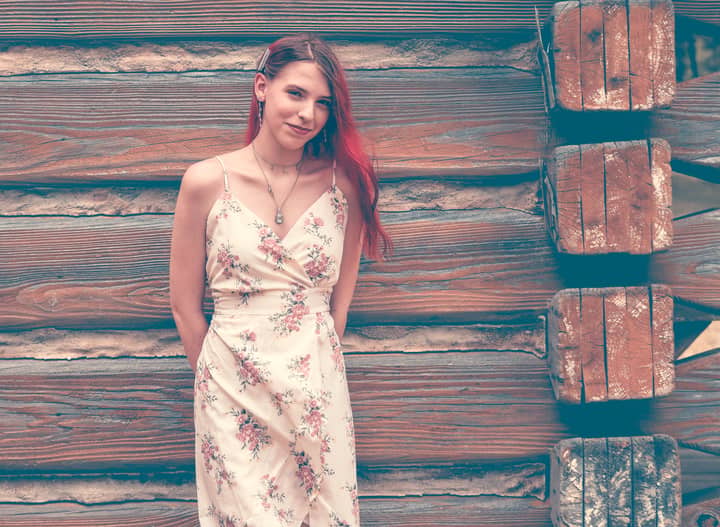 A young woman with red hair poses against a textured wooden wall, wearing a floral dress.