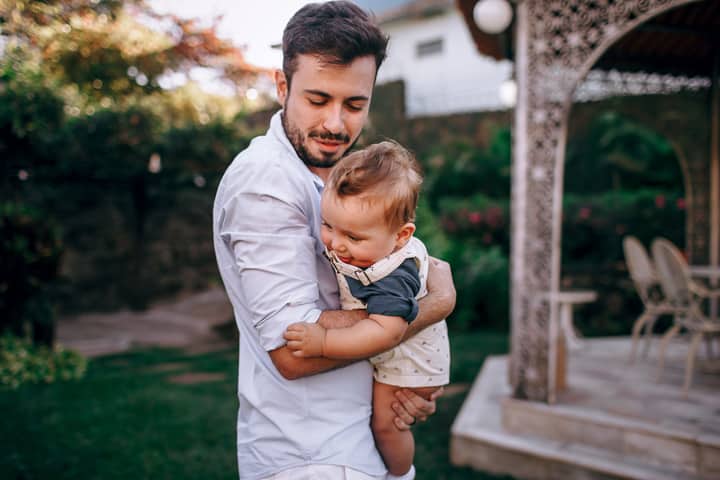 A father lovingly holds his baby in a lush outdoor garden setting.