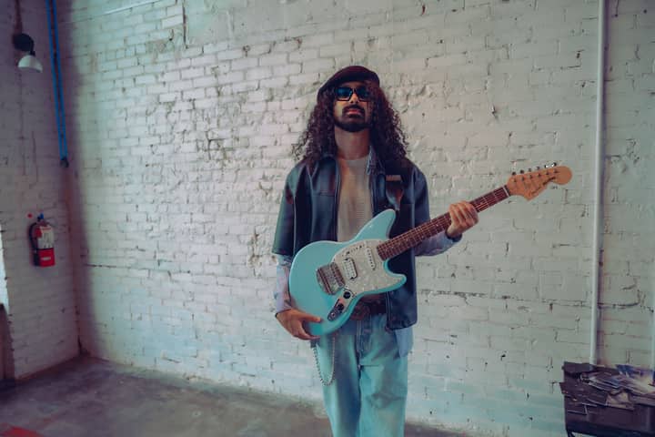 A fashionable musician with sunglasses and a blue electric guitar against a brick wall.