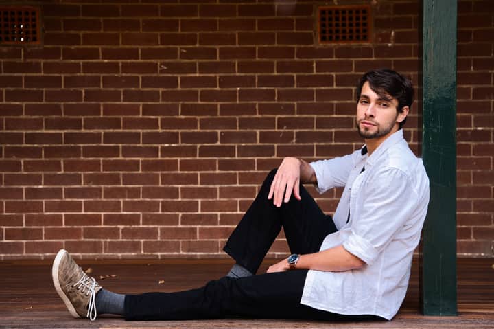 Stylish young man in Sydney sitting against a brick wall, relaxed and fashionable.