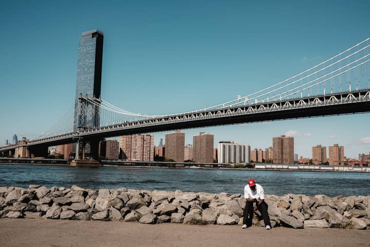 Manhattan Bridge spans over East River with New York City skyline backdrop and person in the foreground.