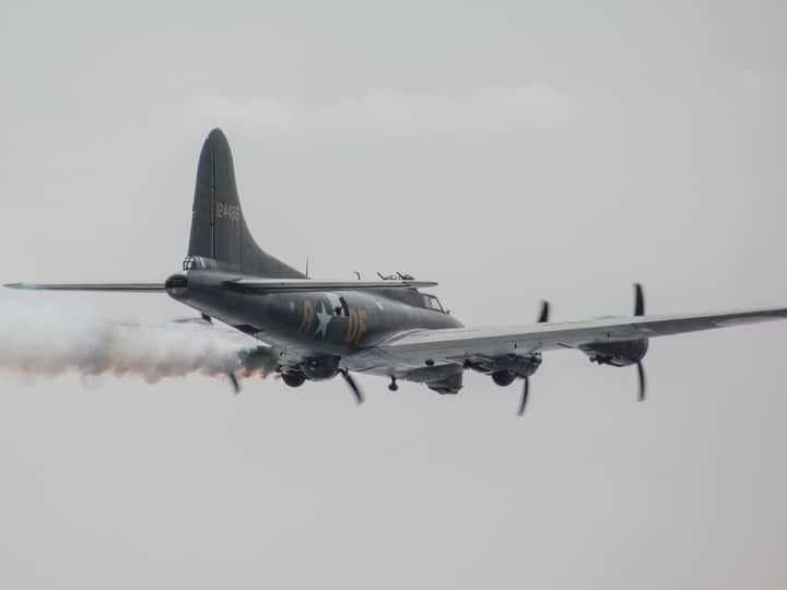 A classic military aircraft flying with a visible smoke trail during daytime.