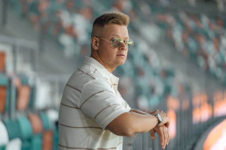 Man in white shirt wearing sunglasses, standing at an outdoor stadium with blurred seating in the background.