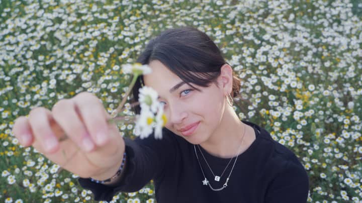 A young woman holds a daisy in a field of flowers, smiling softly.