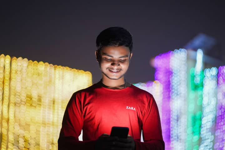 Smiling young man in red shirt using smartphone against a backdrop of festive lights at night.