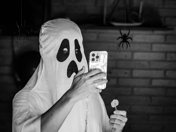 A person in a ghost costume takes a selfie with a smartphone indoors.