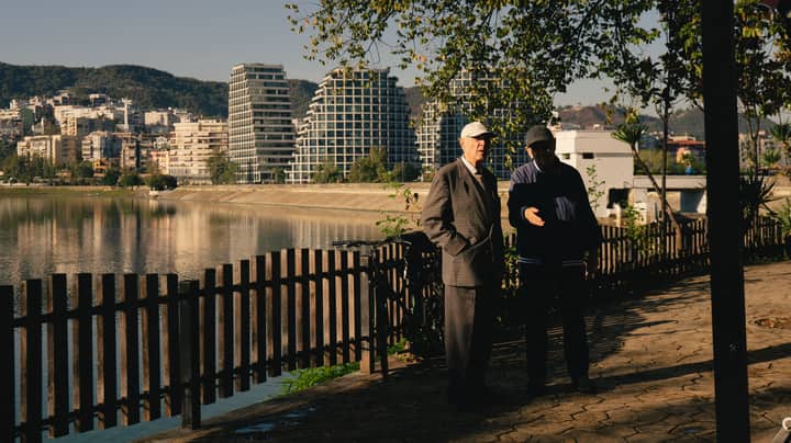 Two senior men engage in conversation along the serene Tirana lakefront, shadowed by modern architecture.