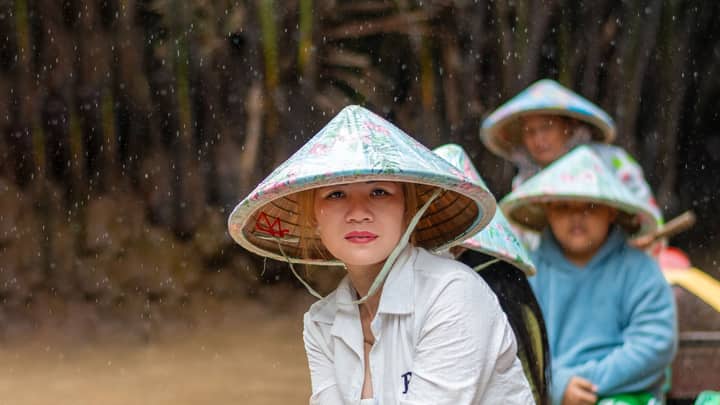 A scene of people wearing traditional hats during a rainy boat ride in Mỹ Tho, Vietnam.