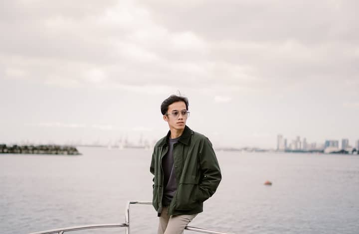 Young man in green jacket aboard a boat with city skyline in background.