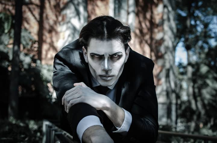 A dramatic portrait of a person with pale makeup wearing a black suit in an outdoor setting.