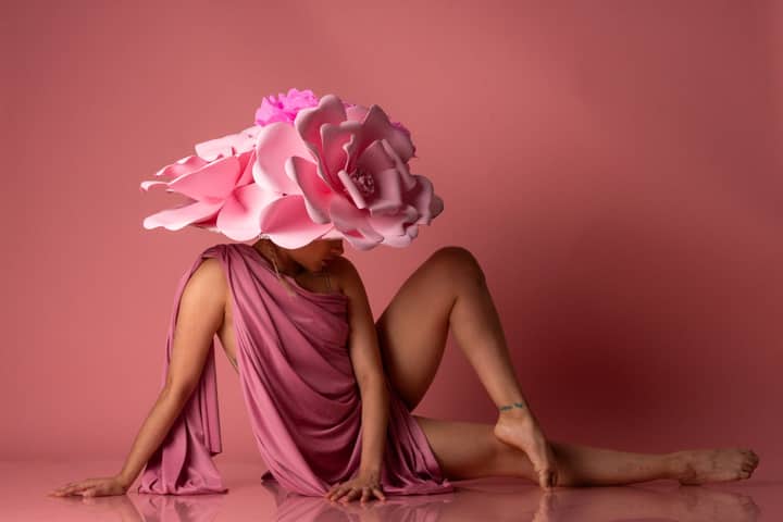 Artistic portrait of a female dancer in pink attire and a large floral hat, set against a pink backdrop.
