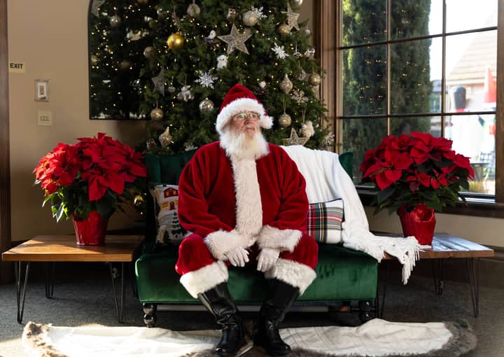 Santa Claus seated on a green couch surrounded by Christmas decor and poinsettia plants.