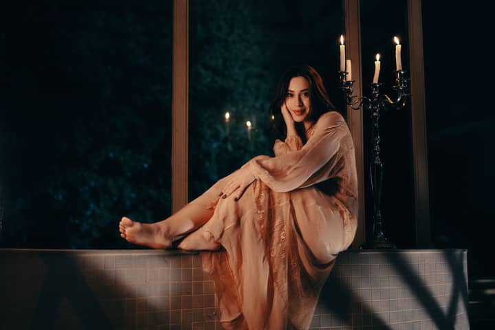 A serene portrait of a woman in a nightgown, seated beside candles, exuding elegance and calm.