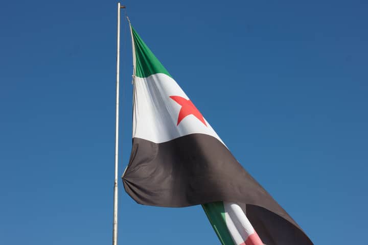Syrian flag with red star waving on a pole against a clear blue sky.