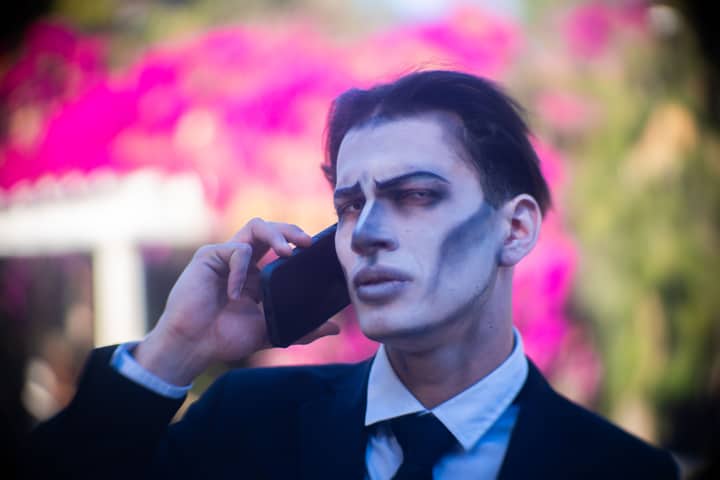 Young man in gothic-style makeup using a smartphone outdoors, surrounded by vibrant flowers.