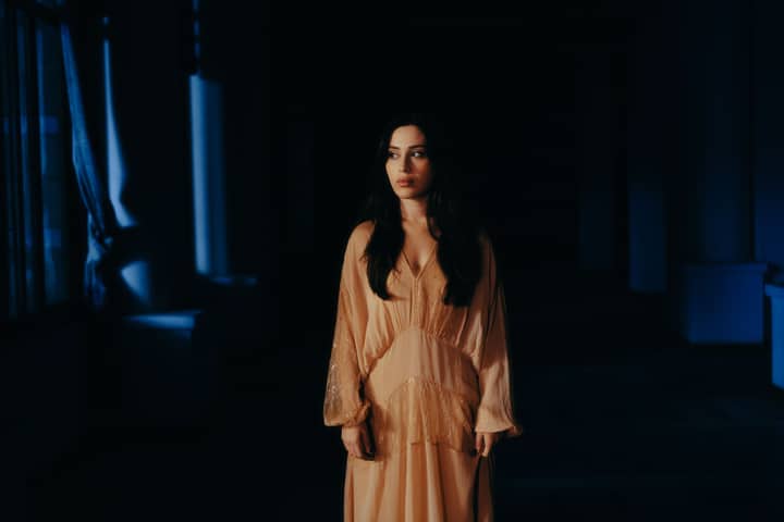 Woman in a flowing dress stands by a window in dramatic lighting.