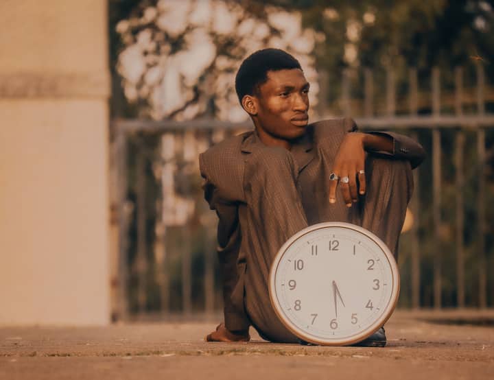 A contemplative man in a suit sits outdoors holding a large clock, embodying the passage of time.