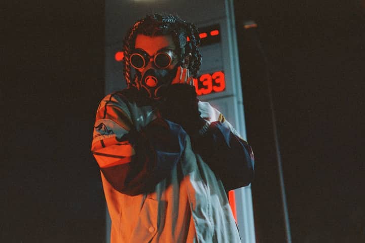 A person in a dramatic gas mask and jacket stands near a lit structure at night.