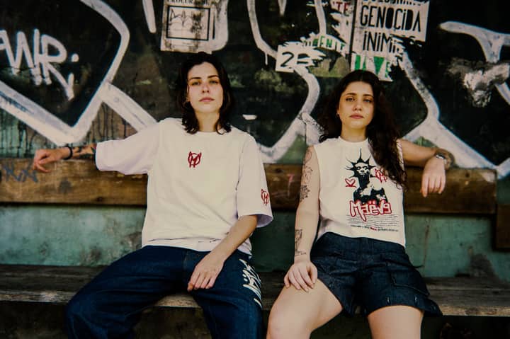 Two young adults in streetwear sit against a graffiti-covered backdrop, exuding urban style.